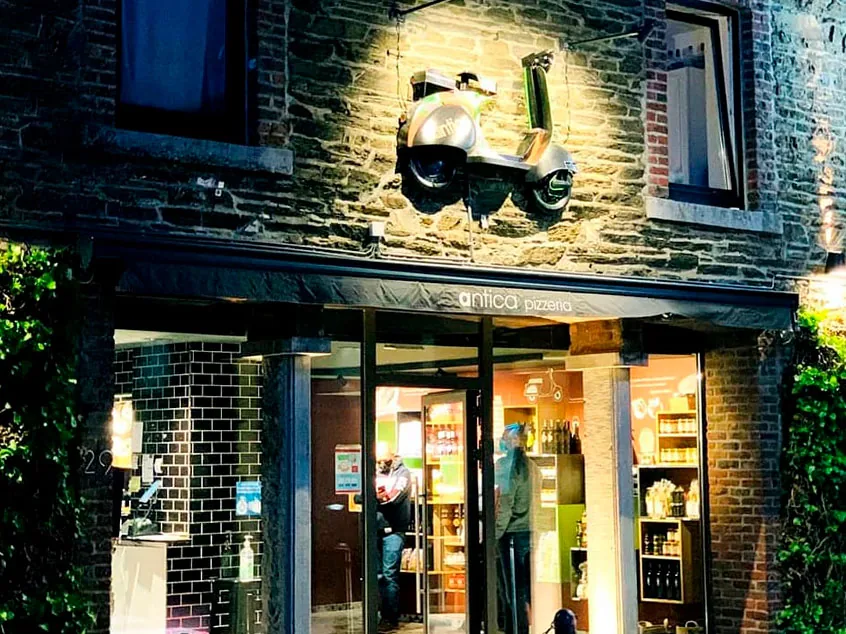 La façade éclairée d'Antica Pizzeria, un soir, avec sa vespa accrochée au mur de briques